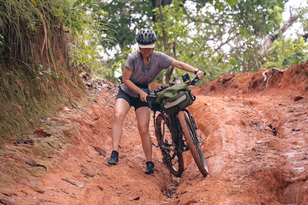 ADVbyCycle Bec walking through the mud with a Road Runner Bags Middle Earth Handlebar Bag in Olive mounted to her mountain bike. The red mud on the trail reflects the light on the background and on the rider.